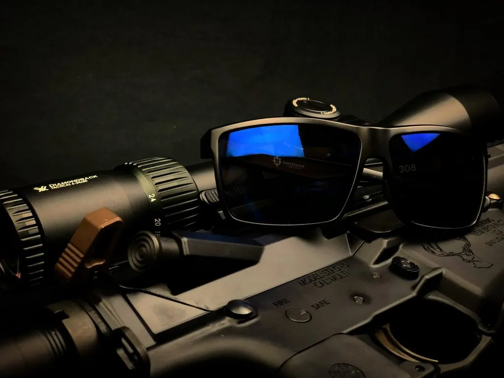 FortKnight Optics 308 polarized sunglasses resting on a matte black rifle with a riflescope, exemplifying the sleek design and sport shooting functionality of the eyewear. The reflective blue lenses highlight the clarity and advanced LightPro Technology by ZEISS, designed for superior visual performance in sport shooting scenarios.