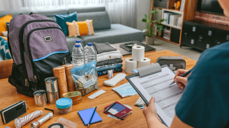 Emergency Preparedness Basics: Navigating the 5 Crucial Phases 5 The image captures a moment of emergency preparedness within a cozy living room setting. A person, seen from behind, is reviewing a detailed emergency plan or checklist clipped to a clipboard. The tabletop is organized with essential emergency supplies: a purple and black backpack, likely prepared and ready to go; bottles of water; canned goods; a plastic bag with dry snacks; a first aid kit; rolled clothing; and various personal items such as a passport, wallet, and a portable radio. The room is warmly lit, suggesting a home environment that is both comfortable and orderly. This scene underscores the importance of being prepared as an individual and contributing to a community-wide culture of readiness for any emergency.