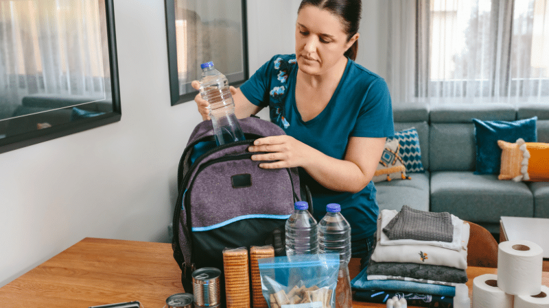 Emergency Preparedness Basics: Navigating the 5 Crucial Phases 3 The image shows a focused woman in a teal top with a floral design on her right shoulder, packing an emergency preparedness kit. She is placing a clear, plastic bottle of water into a dark purple and black backpack. On the wooden table in front of her are more preparedness items: additional bottles of water, a stack of folded clothing, a first aid kit, canned food, and a roll of paper towels. The setting appears to be a well-lit, modern living room with a large window and a comfortable blue couch in the background. This visual serves to guide viewers through the process of assembling a comprehensive emergency preparedness kit.