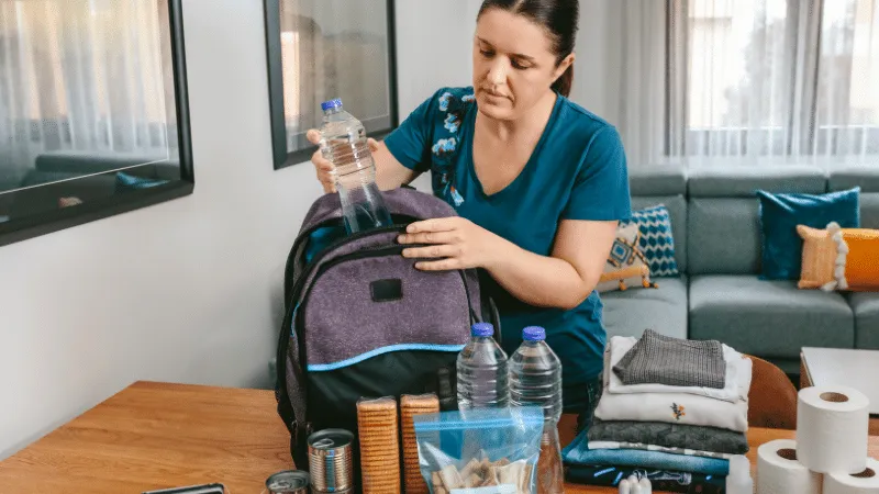 Emergency Preparedness Basics: Navigating the 5 Crucial Phases 3 The image shows a focused woman in a teal top with a floral design on her right shoulder, packing an emergency preparedness kit. She is placing a clear, plastic bottle of water into a dark purple and black backpack. On the wooden table in front of her are more preparedness items: additional bottles of water, a stack of folded clothing, a first aid kit, canned food, and a roll of paper towels. The setting appears to be a well-lit, modern living room with a large window and a comfortable blue couch in the background. This visual serves to guide viewers through the process of assembling a comprehensive emergency preparedness kit.