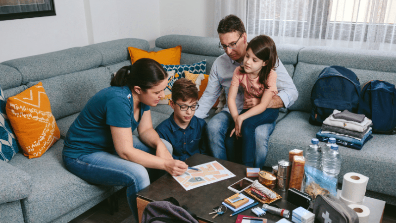 Emergency Preparedness Basics: Navigating the 5 Crucial Phases 4 The image depicts a family of four engaged in emergency planning within their home. A mother, in a blue T-shirt, is pointing to a document on the table, which appears to be an emergency plan, while her son, wearing glasses and a blue shirt, looks on attentively. To the right, a father, wearing glasses and a grey shirt, embraces his young daughter, who is wearing a pink top. They are seated around a coffee table with various emergency supplies laid out, including bottled water, canned food, a first aid kit, toiletries, and a flashlight. Backpacks are also packed and ready, indicating their preparedness for a potential emergency situation. The setting suggests a comfortable living room with a grey couch adorned with colourful pillows. The atmosphere is one of calm and focused preparation.