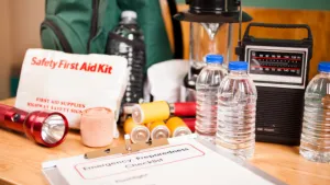 A comprehensive display of emergency preparedness basics laid out on a wooden surface, featuring essential items for crisis situations. Visible items include a 'Safety First Aid Kit' with a clear label, batteries, rolls of adhesive bandages, bottled water, a sturdy flashlight, and a portable radio for emergency broadcasts. In the foreground, there is an 'Emergency Preparedness Checklist' ensuring readiness for various scenarios, emphasizing the importance of preparedness and safety.