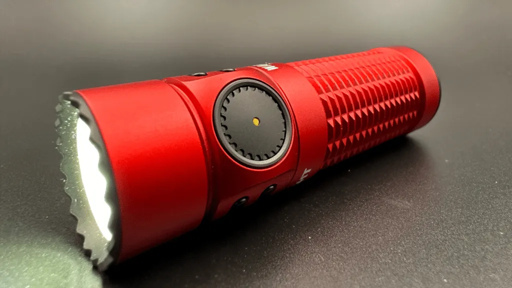 This image presents a close-up view of the side switch on the Olight Warrior Nano flashlight. The flashlight is positioned horizontally with the side switch in clear focus, featuring a black, textured surface for easy operation and a small golden dot at its center, indicative of the LED indicator beneath. The surrounding body of the flashlight is a vivid red, with a pronounced texture along its length for a secure grip. The head of the flashlight has a crenelated bezel, providing a sense of its tactical design. The angle of the photo captures the sleek, compact form of the flashlight, emphasizing its sturdy build and portability. The background is a dark grey, ensuring the details of the flashlight stand out sharply.