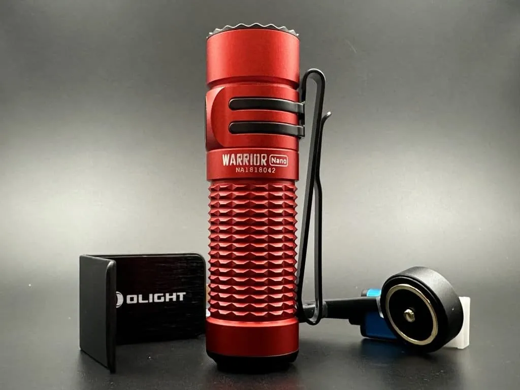 This image features a close-up view of the Olight Warrior Nano flashlight in a striking red coluor. The flashlight is positioned upright, showcasing its textured grip and the name 'WARRIOR Nano' engraved on its side. To the left of the flashlight is a compact, black rectangular Olight branded charger with a USB port visible on its side. To the right is the flashlight's charging cable with a round magnetic charging head in black, accented with a blue rim, connected to a white USB plug. The entire assembly is set against a dark grey background that contrasts with the vibrant red of the flashlight, highlighting the device's rugged design and portability.
