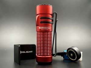 This image features a close-up view of the Olight Warrior Nano flashlight in a striking red coluor. The flashlight is positioned upright, showcasing its textured grip and the name 'WARRIOR Nano' engraved on its side. To the left of the flashlight is a compact, black rectangular Olight branded charger with a USB port visible on its side. To the right is the flashlight's charging cable with a round magnetic charging head in black, accented with a blue rim, connected to a white USB plug. The entire assembly is set against a dark grey background that contrasts with the vibrant red of the flashlight, highlighting the device's rugged design and portability.