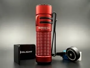 This image features a close-up view of the Olight Warrior Nano flashlight in a striking red coluor. The flashlight is positioned upright, showcasing its textured grip and the name 'WARRIOR Nano' engraved on its side. To the left of the flashlight is a compact, black rectangular Olight branded charger with a USB port visible on its side. To the right is the flashlight's charging cable with a round magnetic charging head in black, accented with a blue rim, connected to a white USB plug. The entire assembly is set against a dark grey background that contrasts with the vibrant red of the flashlight, highlighting the device's rugged design and portability.