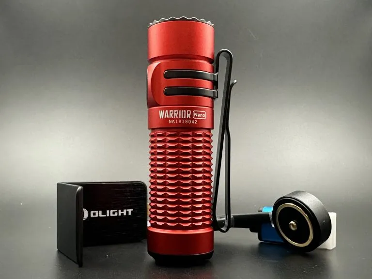 This image features a close-up view of the Olight Warrior Nano flashlight in a striking red coluor. The flashlight is positioned upright, showcasing its textured grip and the name 'WARRIOR Nano' engraved on its side. To the left of the flashlight is a compact, black rectangular Olight branded charger with a USB port visible on its side. To the right is the flashlight's charging cable with a round magnetic charging head in black, accented with a blue rim, connected to a white USB plug. The entire assembly is set against a dark grey background that contrasts with the vibrant red of the flashlight, highlighting the device's rugged design and portability.