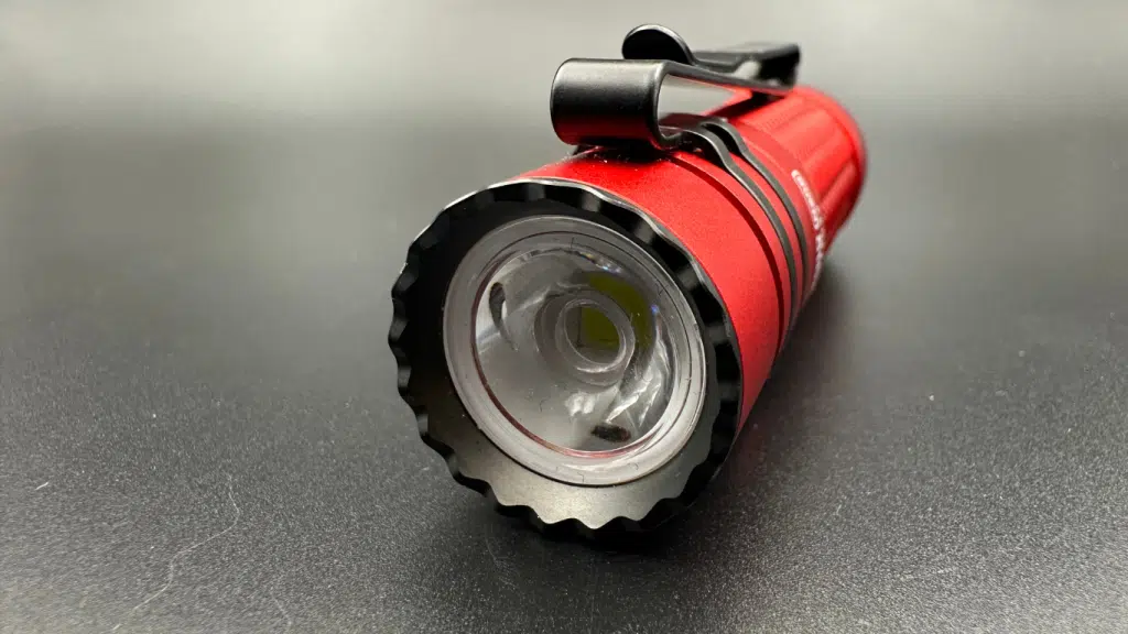 This image displays a close-up view of the front end of the Olight Warrior Nano flashlight, emphasizing its aggressive bezel and LED light. The flashlight is shown at an angle that reveals the serrated design of the bezel, intended for tactical use, with each tooth-like point reflecting the light differently, creating a pattern of highlights and shadows. The central LED is visible as a small yellow rectangle set deep within a clear, smooth reflector, which magnifies the LED's intensity. A sturdy clip arcs over the top of the flashlight, providing a secure attachment point for carrying. The body of the flashlight is coated in a bright red finish that contrasts with the black features, such as the bezel and clip. The grey background serves to focus attention on the flashlight's details, illustrating its potential for both everyday use and emergency situations.