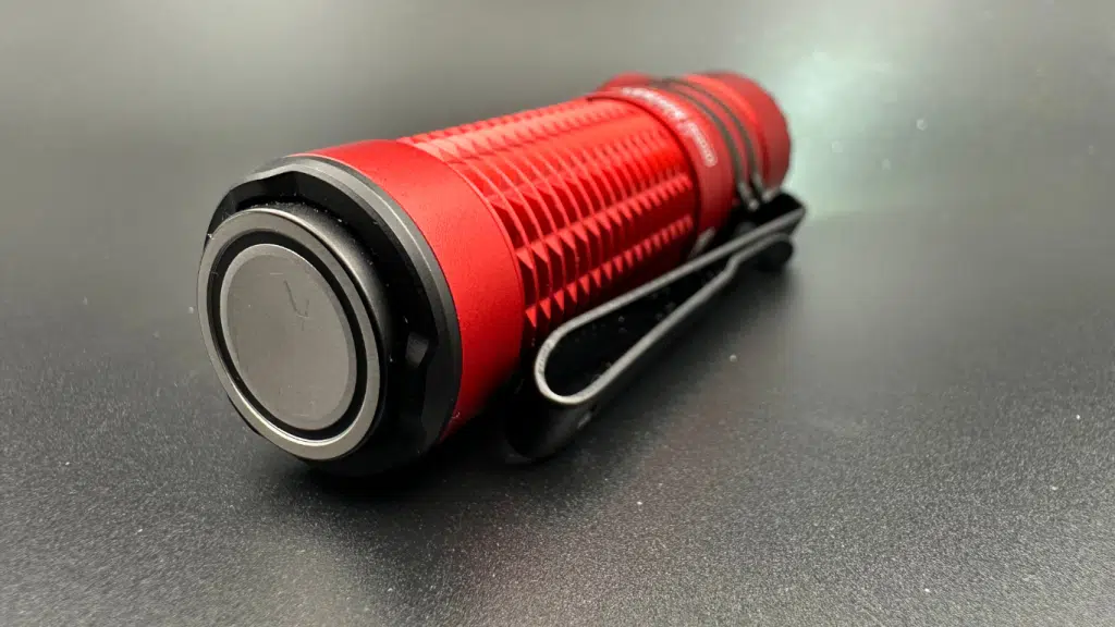 This image displays the tail switch of the Olight Warrior Nano flashlight, offering a detailed view of its design and features. The flashlight is oriented diagonally across the frame, highlighting the large, prominent black tail switch, which contrasts with the red body of the device. The switch has a slightly raised, smooth surface for easy pressing, encircled by a silver-colored edge that adds to its sleek appearance. A sturdy, black dual-purpose clip attached to the side of the flashlight provides versatility for carrying and mounting. The body of the flashlight is ribbed for grip and the red color is vivid against the dark grey, almost black, background. The image clearly demonstrates the robust, tactical design of the tail switch, which is an integral part of the flashlight's functionality.